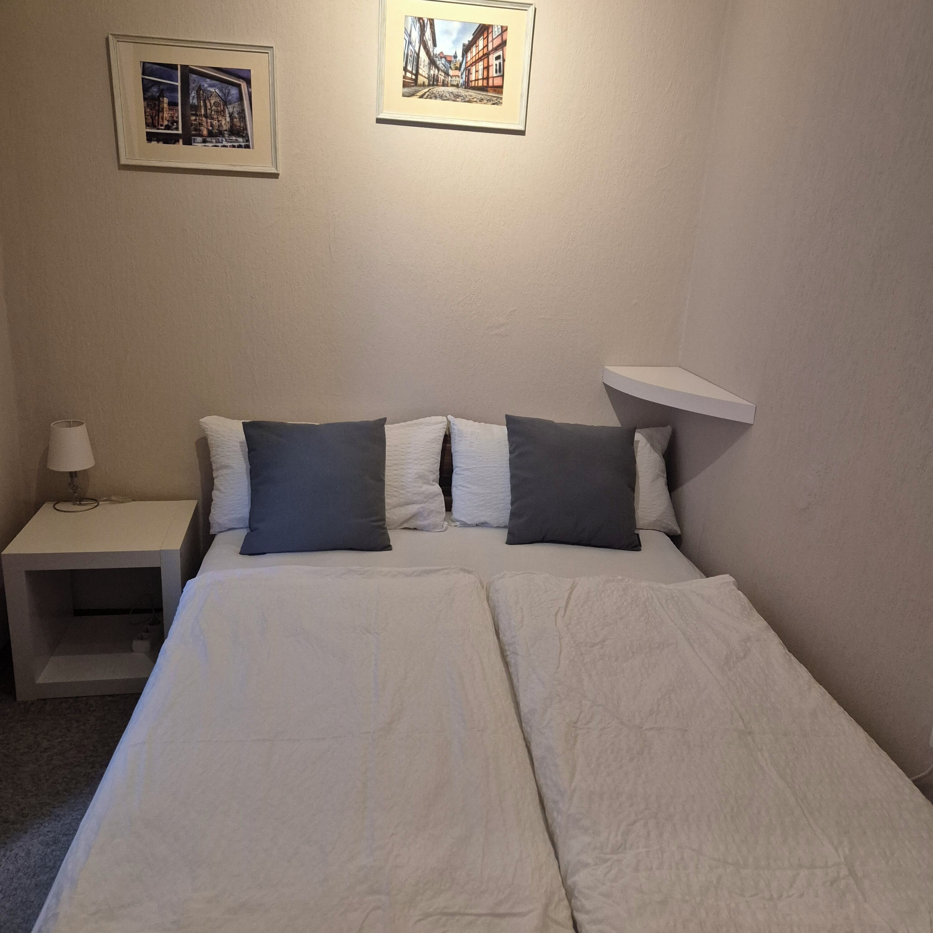 Un lit double avec une literie blanche et deux oreillers, une table de chevet avec une lampe, et deux tableaux encadrés accrochés au mur.