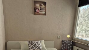 A neatly made bed with a patterned pillow and a rolled towel, a bedside table with a lamp and a clock, and a wall-mounted picture of stones and a flower. - Vacation apartment "Amina" in Hahnenklee (Goslar)