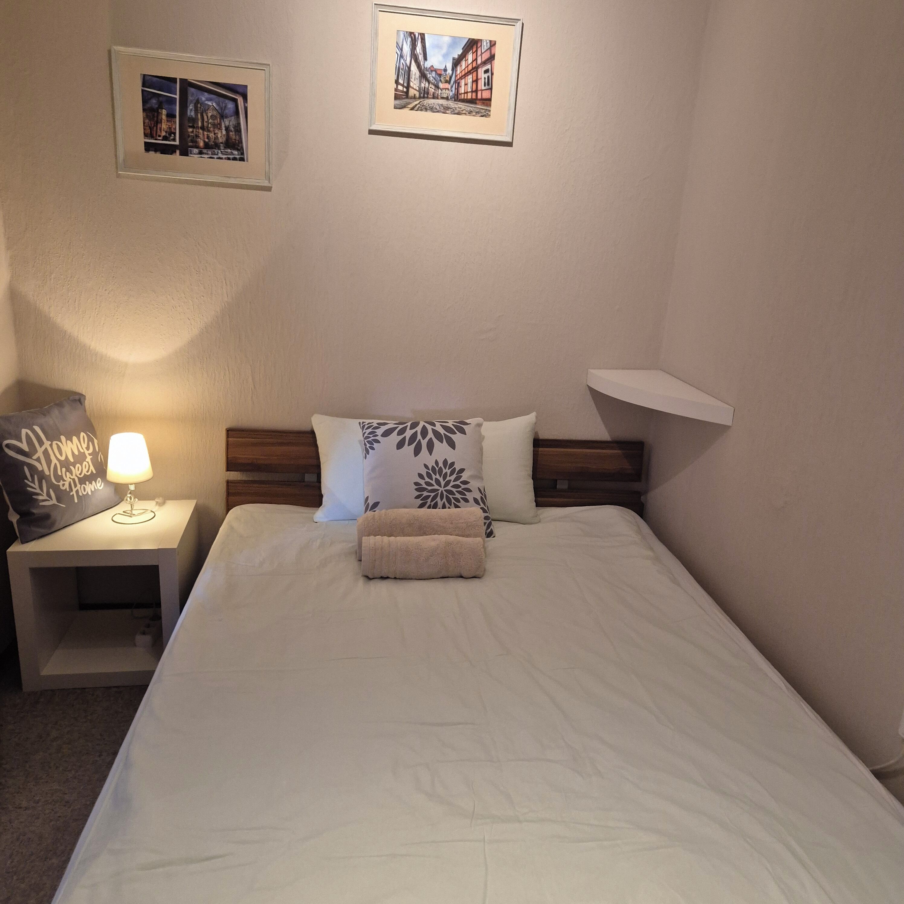 Een net opgemaakt bed met een witte dekbedovertrek en een opgevouwen handdoek, een bedlampje, een nachtkastje met een decoratief kussen, en twee ingelijste foto's aan de muur.