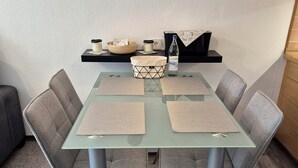 A dining table with four grey chairs, a glass top, and woven placemats. There is a black shelf above the table holding candles, a bottle, and a bowl. - Vacation apartment "Amina" in Hahnenklee (Goslar)