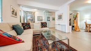 Una sala de estar moderna con un sofá, una mesa de centro de vidrio y alfombras coloridas. Hay cuadros enmarcados en las paredes y una escultura de madera en la esquina.