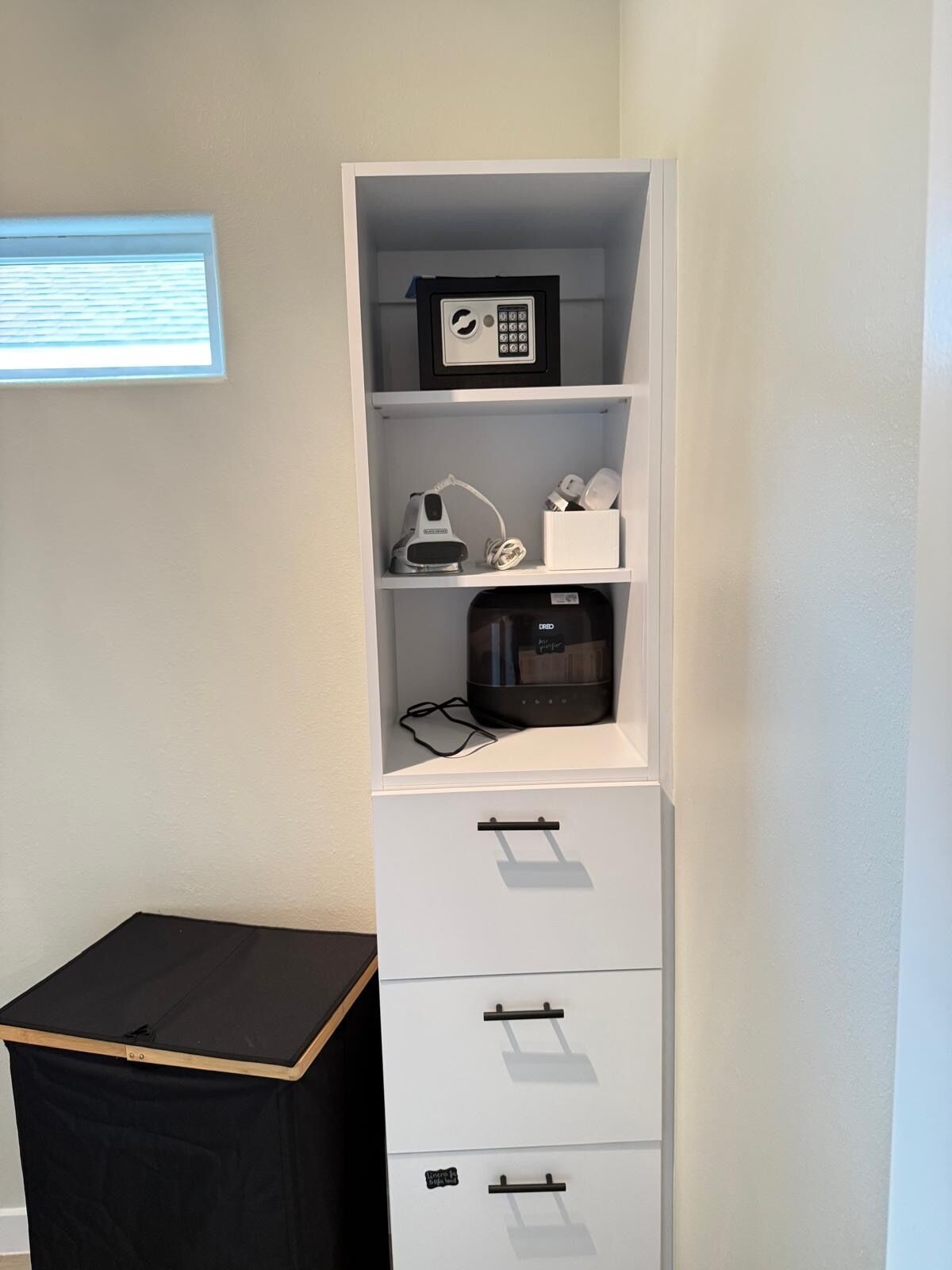 Un meuble de rangement blanc, doté de tiroirs et d’étagères, contenant un téléphone, une machine à café et divers petits objets.