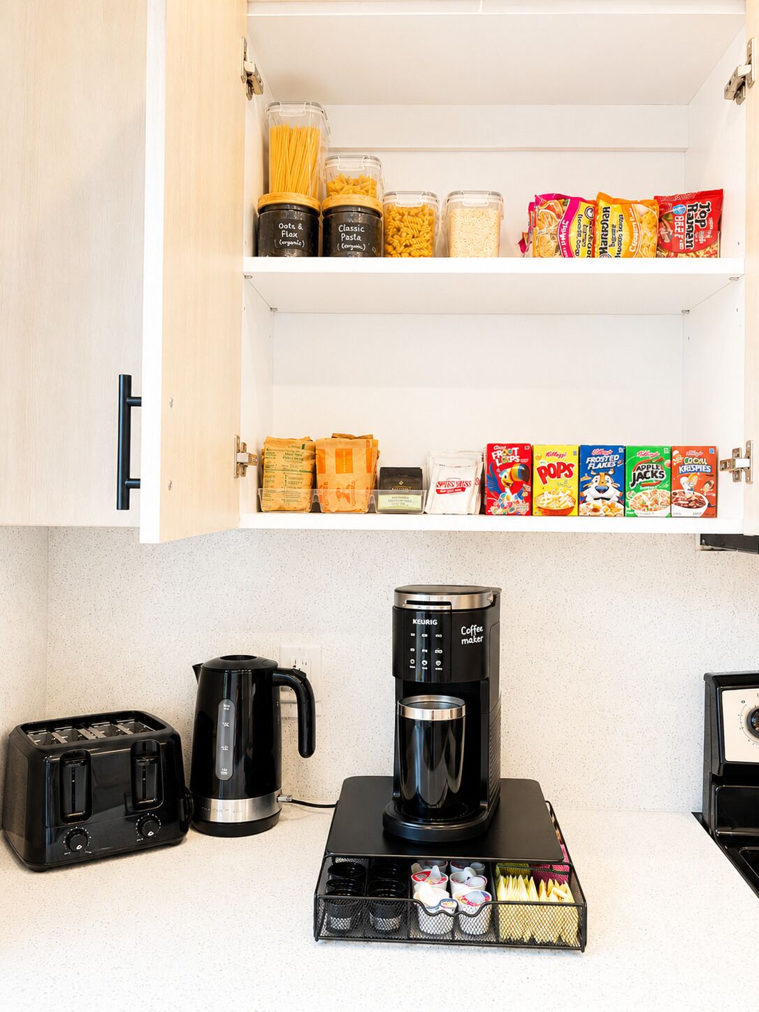 Et køkken med kaffemaskine, brødrister og diverse fødevarer på hylder.