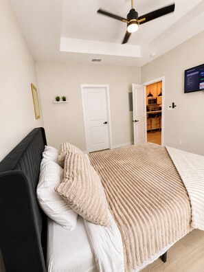 Une chambre à coucher comprenant un lit, un ventilateur de plafond, un téléviseur et une porte donnant sur la cuisine.