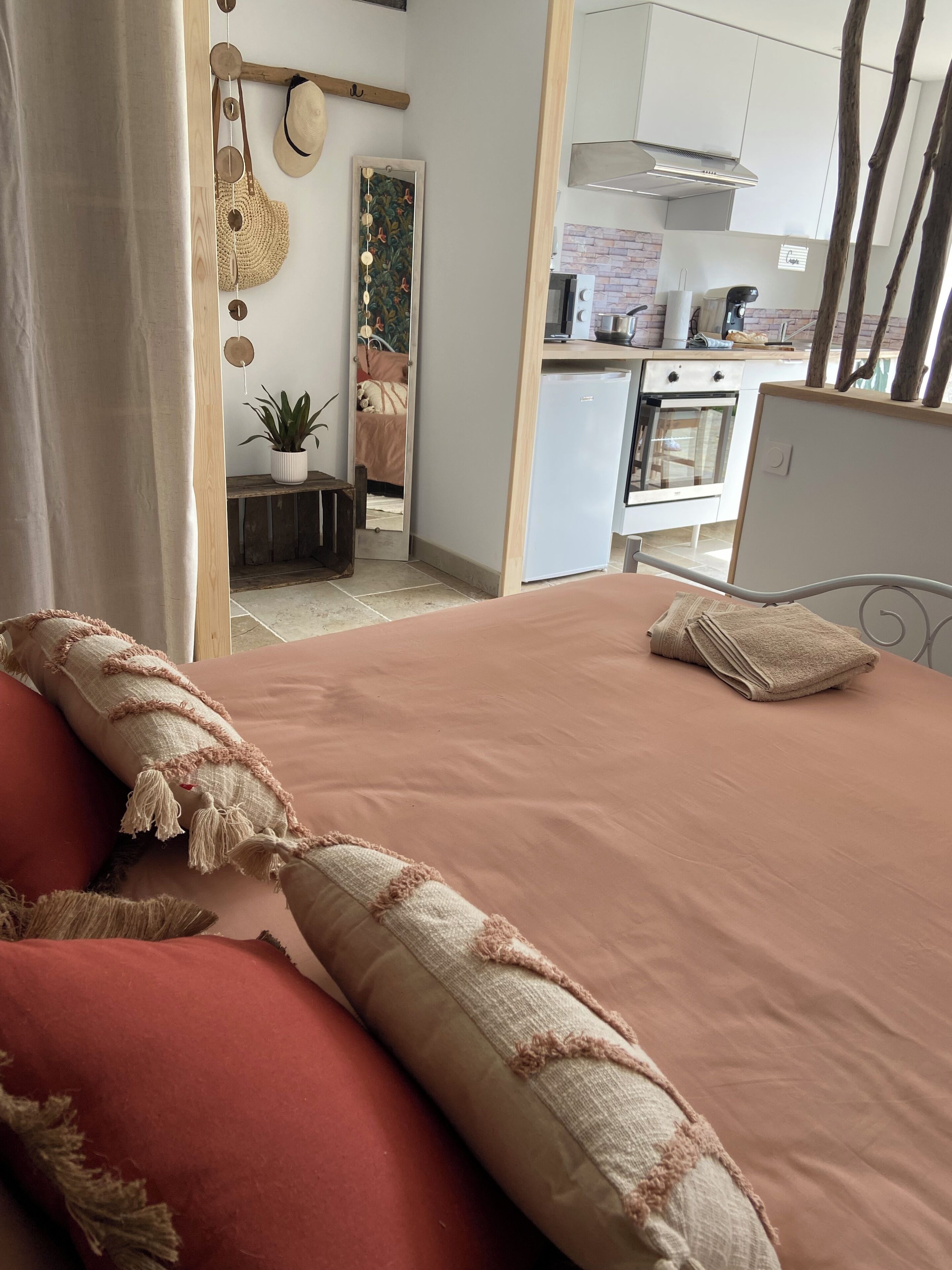 A bedroom with a bed, pillows, a bedside table, a mirror, and a view into a kitchen area.