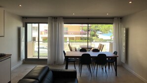 A modern living room with a sofa, dining table, and chairs, large windows with curtains, and a view of buildings and greenery outside. - ARCACHON Eyrac Front de mer Apartment 68M2 with large balcony (Arcachon)
