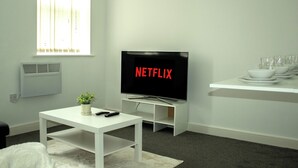 Una sala de estar moderna con un televisor de pantalla plana mostrando “NETFLIX”, una mesa de centro blanca con una planta, un control remoto y un sofá blanco.