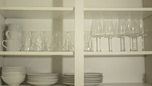 Una estantería blanca con cuencos y platos blancos apilados, y vasos de plástico transparente y copas de vino.