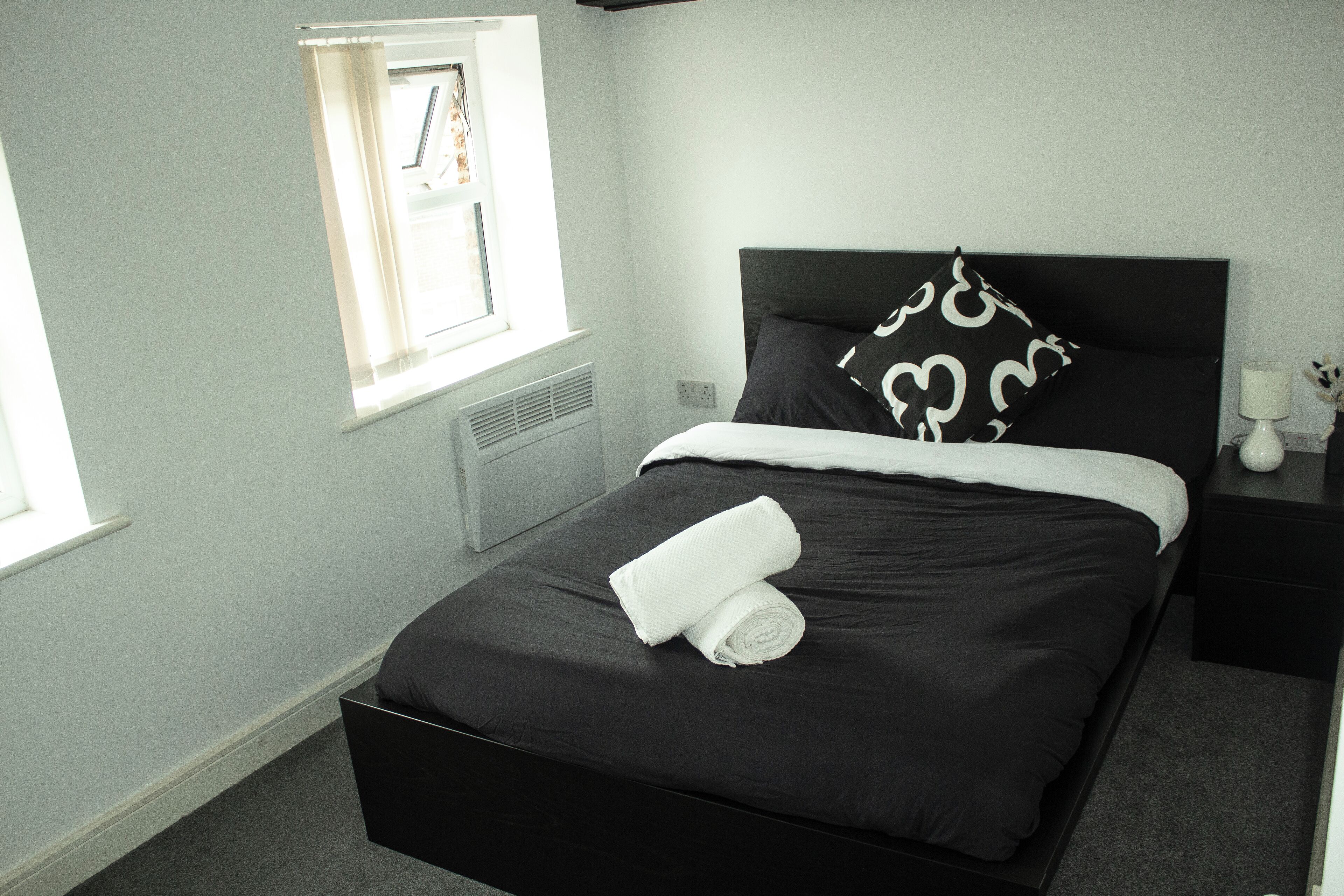 A neatly made bed with a black headboard, a window with blinds, and a bedside table with a lamp.