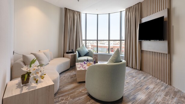 A modern hotel room with a curved sofa, a round armchair, a small coffee table, and a flat-screen TV mounted on the wall. - Amriya Hotel (Madinah)