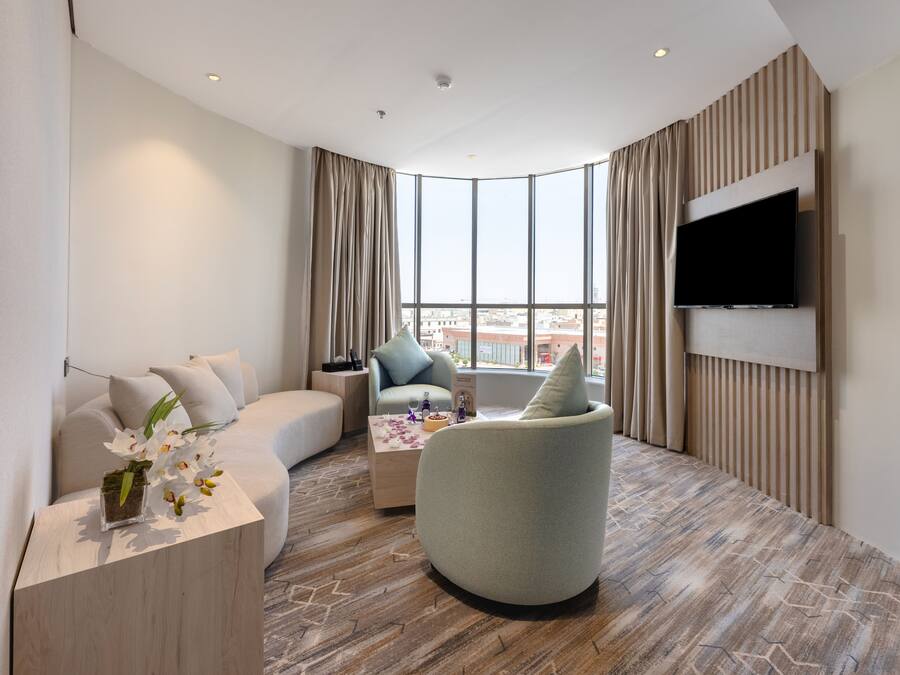 A modern hotel room with a curved sofa, a round armchair, a small coffee table, and a flat-screen TV mounted on the wall.