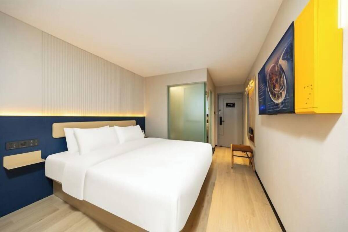 Ein Hotelzimmer mit Bett, Nachttisch, Wandlampe, Tür und Flur.