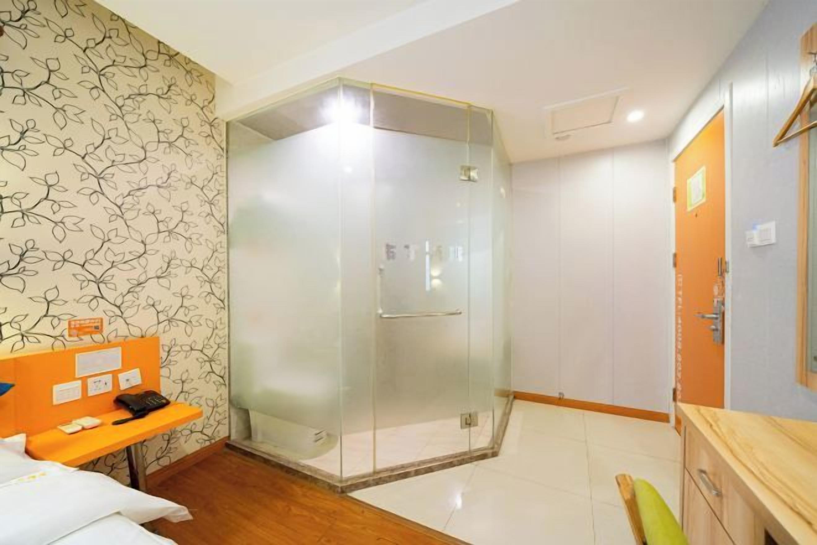 A hotel room with a glass-enclosed shower, a wooden bench, a bed, and a wooden cabinet.