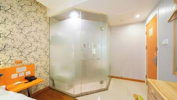 A hotel room with a glass-enclosed shower, a wooden bench, a bed, and a wooden cabinet.