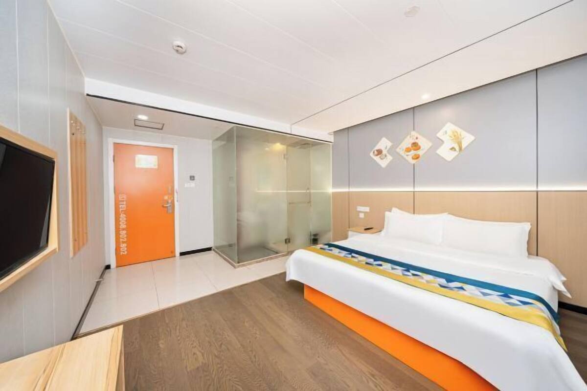 A modern hotel room with a bed, a television, a glass shower enclosure, and a door marked 'HOTEL'.