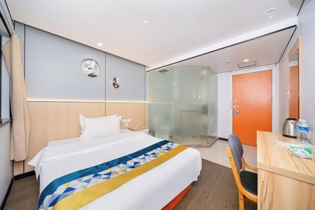 A hotel room with a bed, a desk, a chair, a glass shower enclosure, and a door.