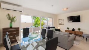 A modern living room with a glass dining table, grey sofa, and a TV mounted on the wall. - Eagle Beach - Luxury Townhome with Private Pool - TH 11 (Noord)
