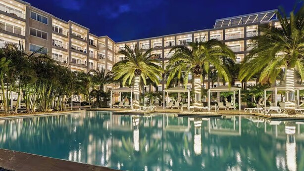 A large indoor swimming pool with lounge chairs and palm trees, surrounded by a multi-story hotel building. - Eagle Beach - Luxury Townhome with Private Pool - TH 11 (Noord)