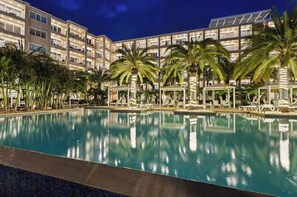 Una amplia piscina interior con sillones reclinables y palmeras, rodeada por un edificio de hotel de varios pisos.