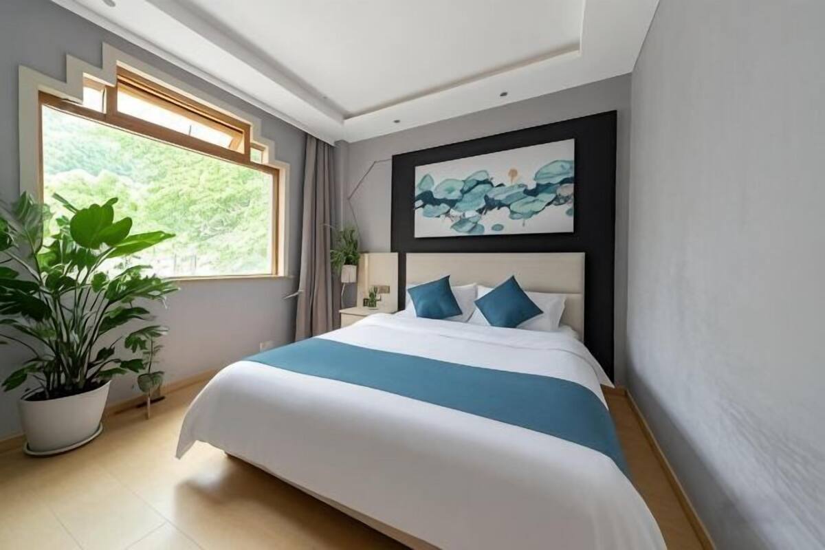 Habitación de hotel moderna con una cama grande, una ventana con vistas a zonas verdes, un cuadro en la pared y una planta en maceta.
