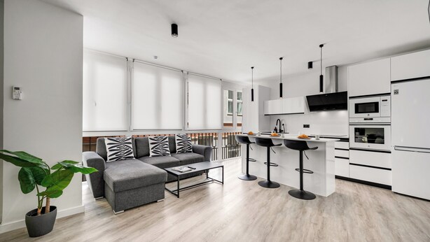 Un appartement moderne avec un canapé gris, une table basse, une cuisine dotée de meubles blancs et un balcon avec une balustrade.