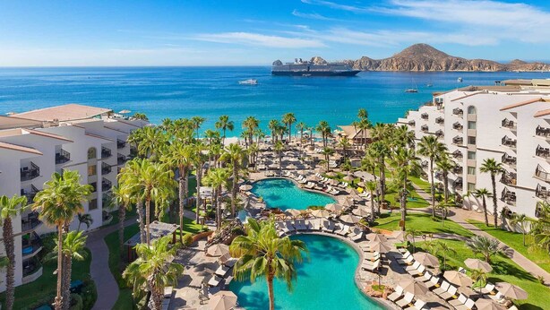 Un resort con piscina central, palmeras y vistas al océano y a un crucero.