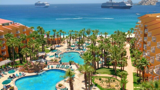 A resort with multiple swimming pools, palm trees, and a view of the ocean with cruise ships.
