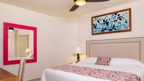 Habitación de hotel con cama, escritorio, silla, ventilador de techo, espejo en la pared, lámpara de noche y un adorno mural.