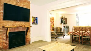 Un salon avec une cheminée en briques, un téléviseur à écran plat fixé au-dessus, et une table basse en bois. La pièce est également équipée de deux fauteuils et d’un canapé.