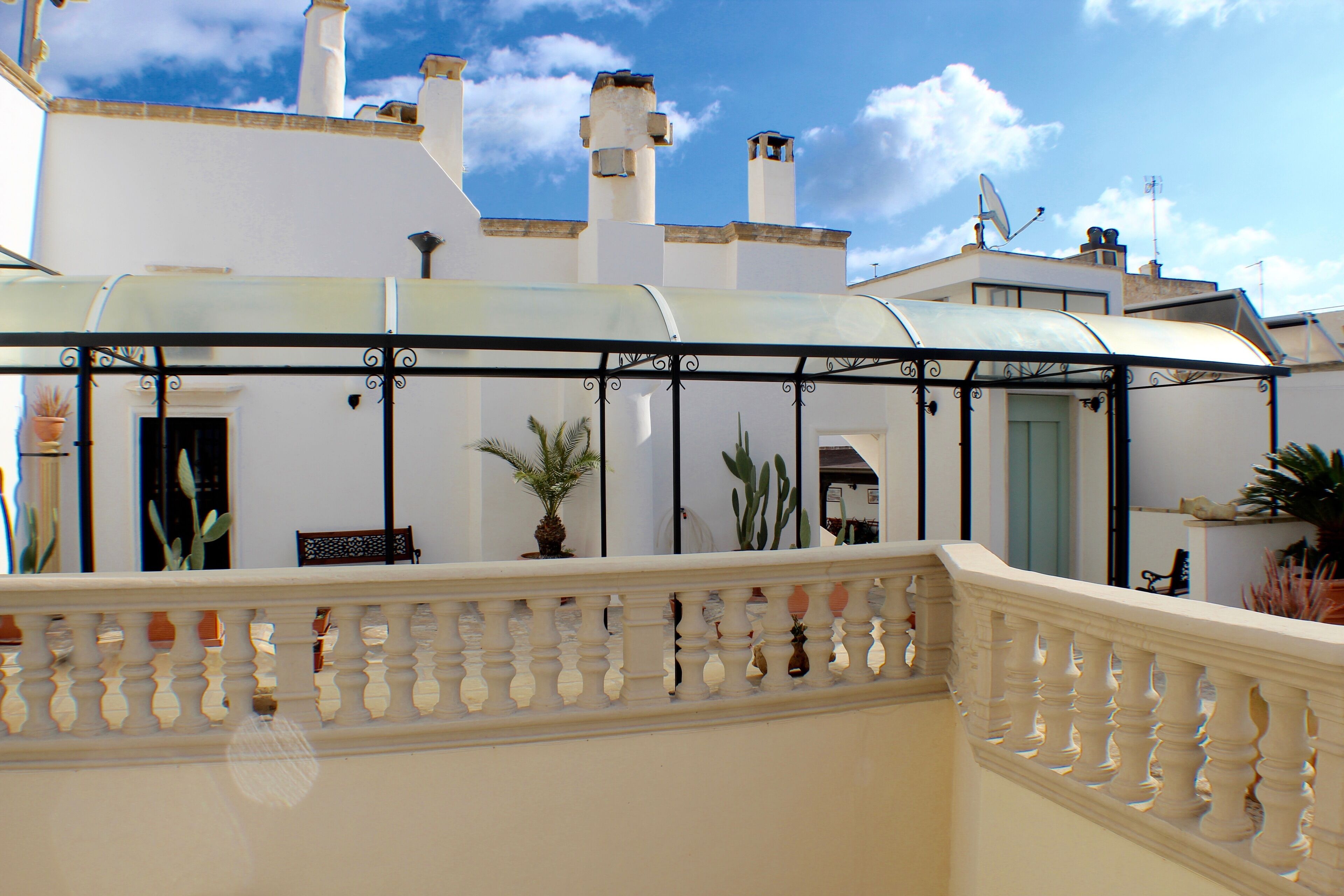 A covered outdoor area with a balustrade, potted plants, and a building with white walls and chimneys.