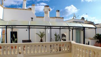 A covered outdoor area with a balustrade, potted plants, and a building with white walls and chimneys.