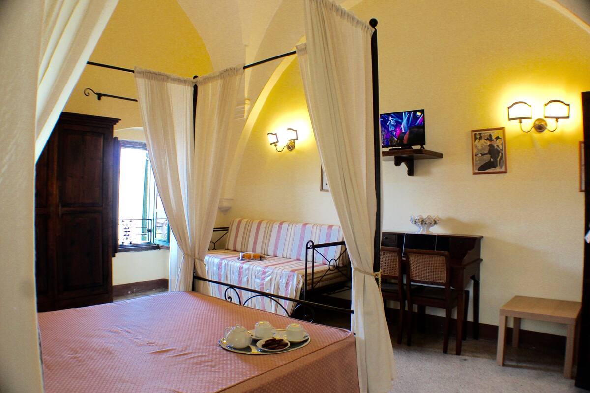 Ein Bett mit Baldachin, ein kleiner Tisch mit einer Teekanne und Tassen, ein Fernseher auf einem Regal und ein Schreibtisch mit Stühlen.