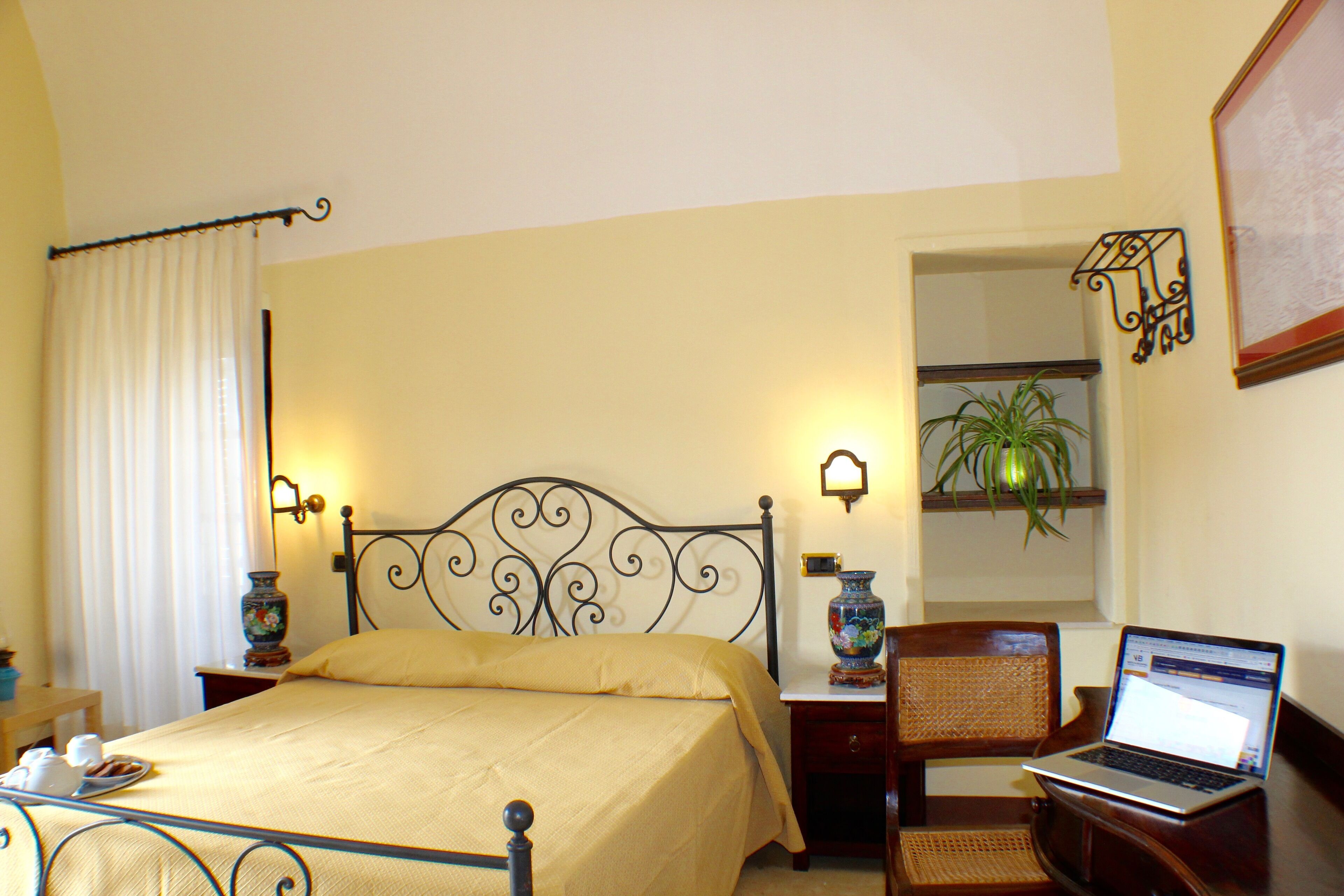 A neatly arranged bedroom with a bed, bedside table, lamp, laptop, and a potted plant.