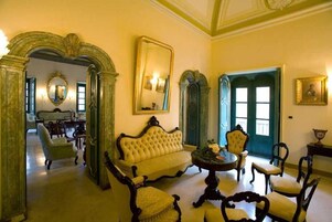 Una habitación con sofá, sillas, una mesa y un espejo. Se ven puertas y una ventana al fondo.