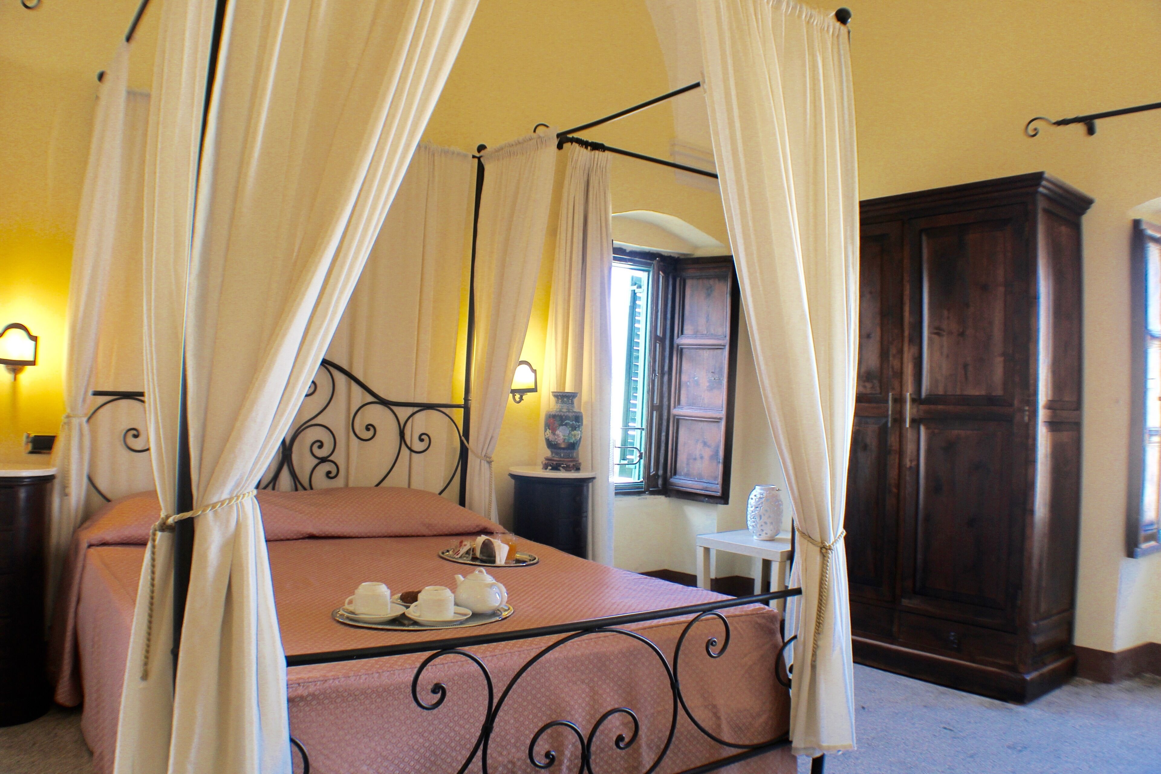 A four-poster bed with a tray of tea cups and saucers, a wooden wardrobe, and a small table with a lamp.