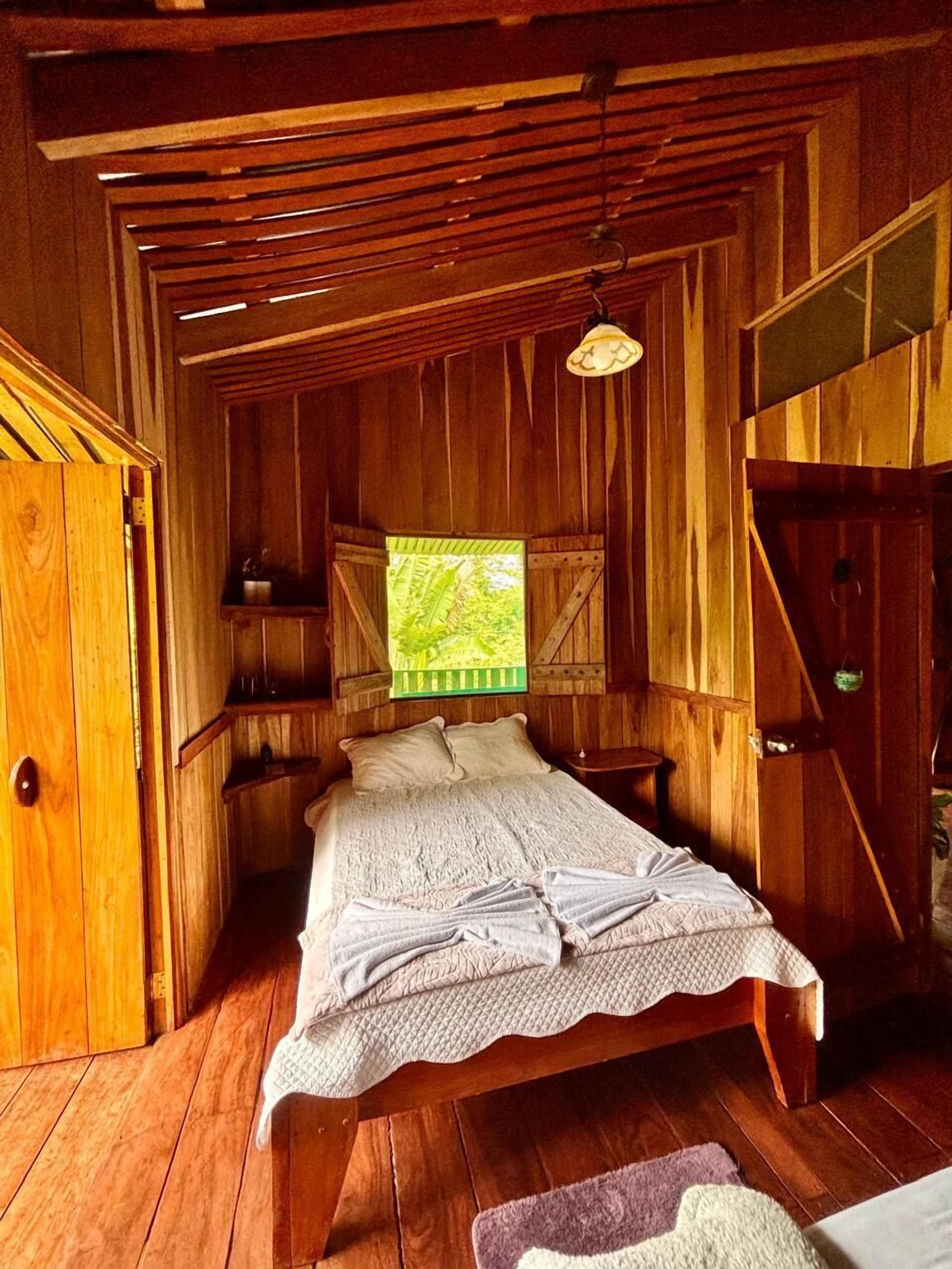 A wooden cabin bedroom with a bed, a window with a view of greenery, and a rustic ceiling.