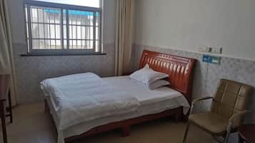 A single bed with a wooden headboard, a chair, a window with curtains, and a wall-mounted light switch.