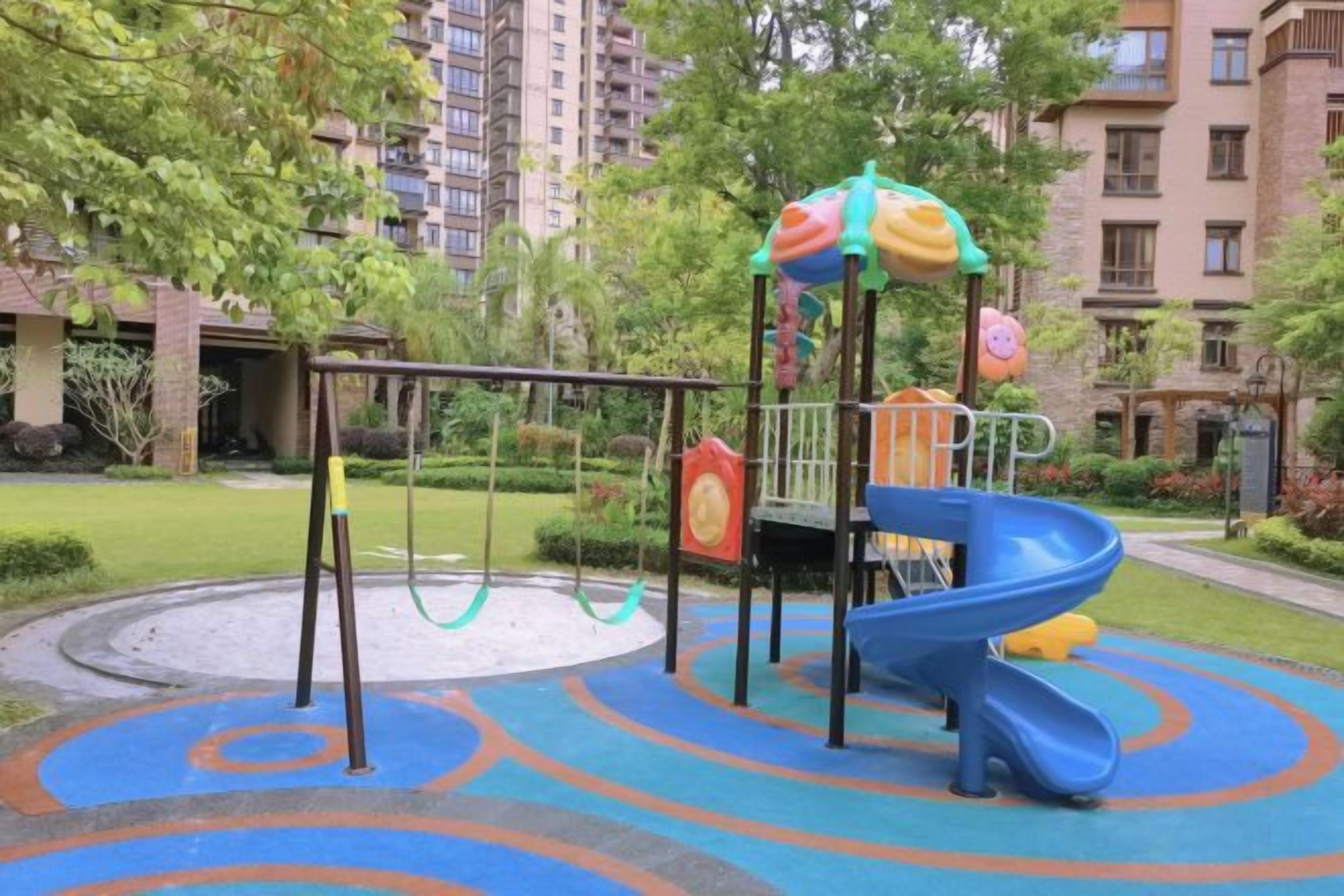 Un parque infantil con tobogán y columpios, rodeado de follaje y edificios residenciales.