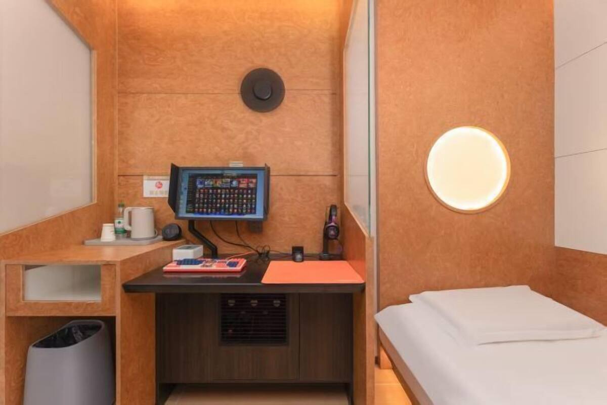 Camera d'albergo compatta con scrivania, letto, cestino e lampada a parete.