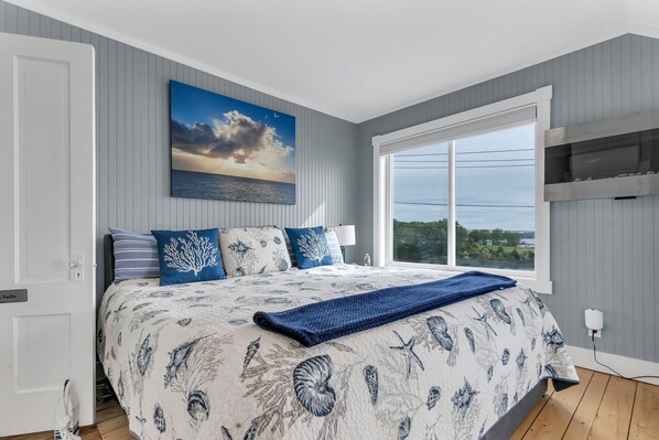 A bedroom with a large bed, a view of the sea, and a painting of a sunset.