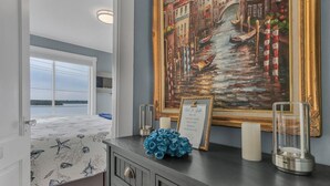 A bedroom with a large window, a bed with a starfish pattern, a painting of a canal scene, and a framed menu on the dresser. - Boutique Luxury Waterfront w/Pool & Sunset Views-Sakonnet Sunset Manor, Tiverton (Tiverton)
