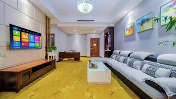 A modern hotel room with a large flat-screen TV mounted on the wall, a sofa, a coffee table, and a wooden bench.