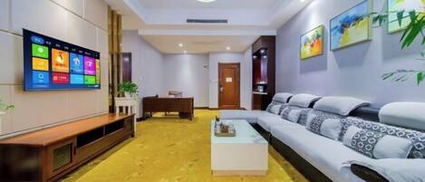 A modern hotel room with a large flat-screen TV mounted on the wall, a sofa, a coffee table, and a wooden bench.
