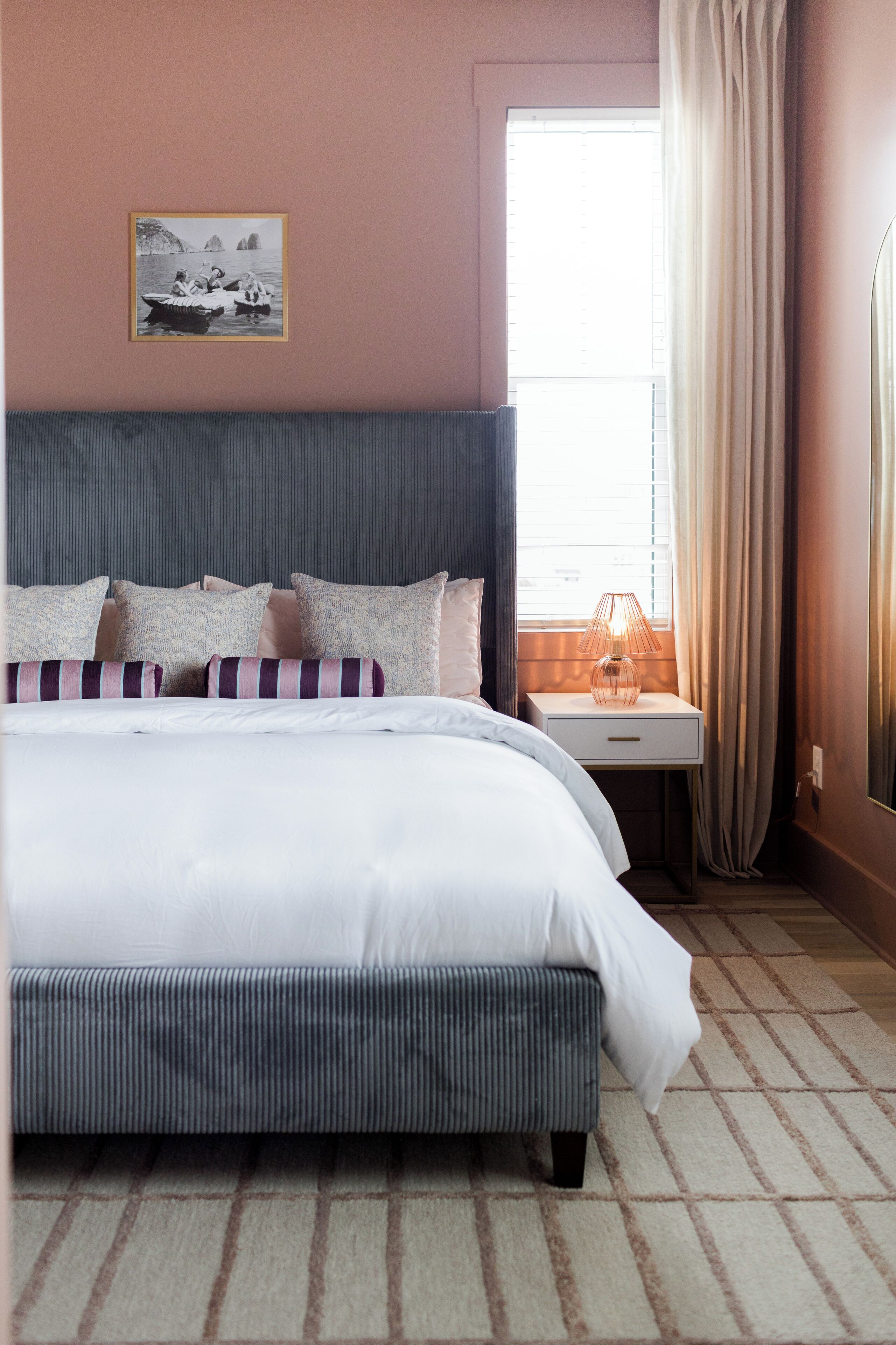 A bedroom with a large bed, a bedside table with a lamp, a framed picture on the wall, and a window with curtains.