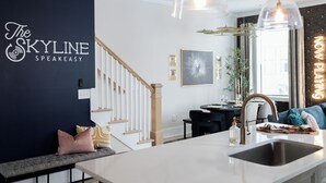 Une cuisine moderne avec un plan de travail blanc, un évier en acier inoxydable et un coin repas comprenant une table ronde et des chaises. On y trouve un escalier avec des rampes blanches et un mur foncé orné d’un logo.