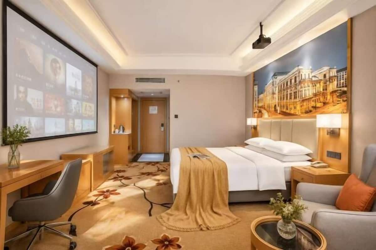 Hotellihuone, jossa on sänky, työpöytä, tuoli ja suuri näyttö, jolla esitetään kuvia.
