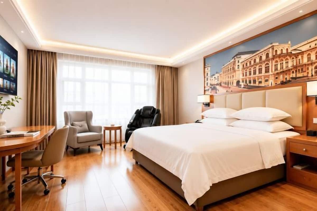 Hotellihuone, jossa on suuri sänky, työpöytä, tuoli, hierontatuoli ja maalaus, jossa on kuvattuna rakennus.