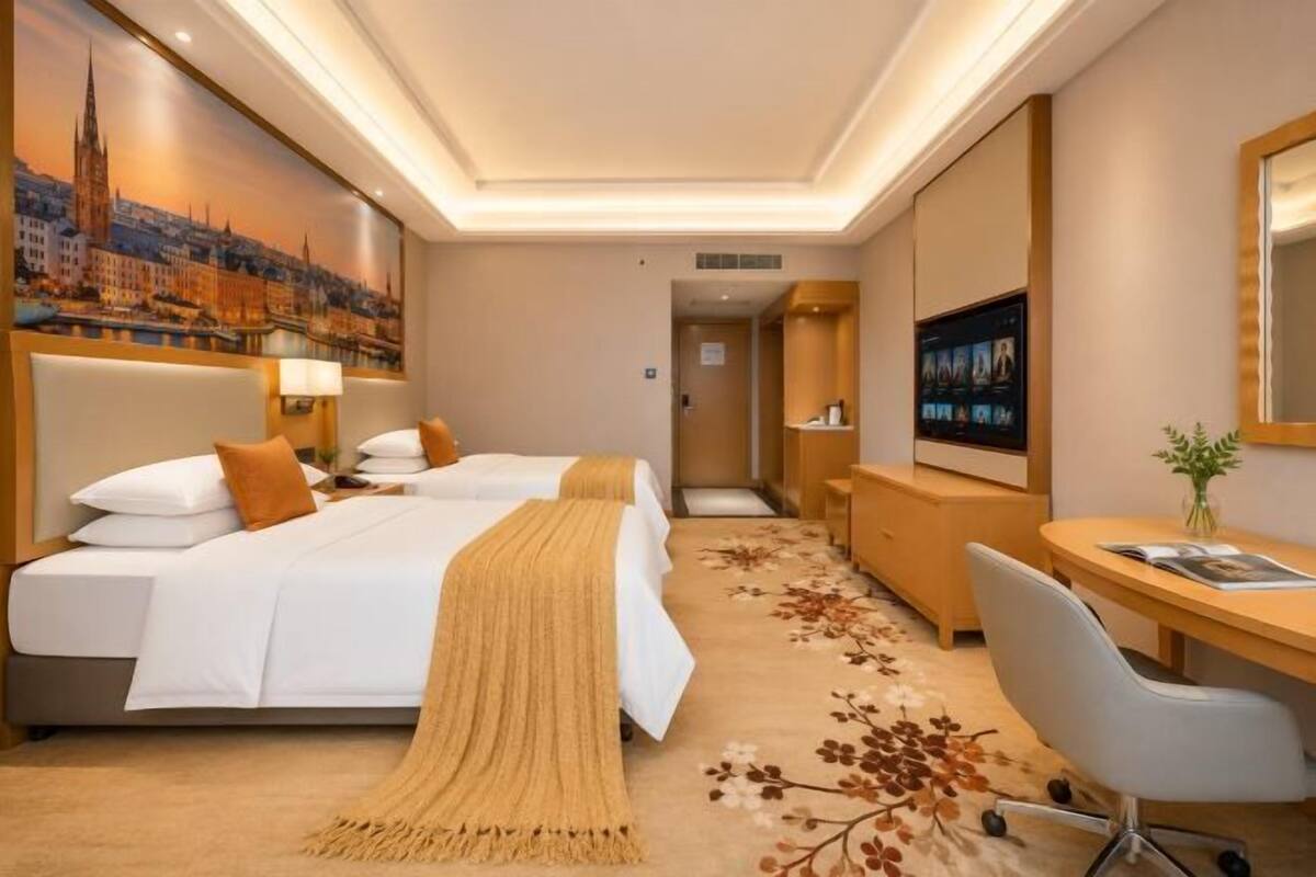 A hotel room with two beds, a desk, a chair, a TV, and a painting of a cityscape.
