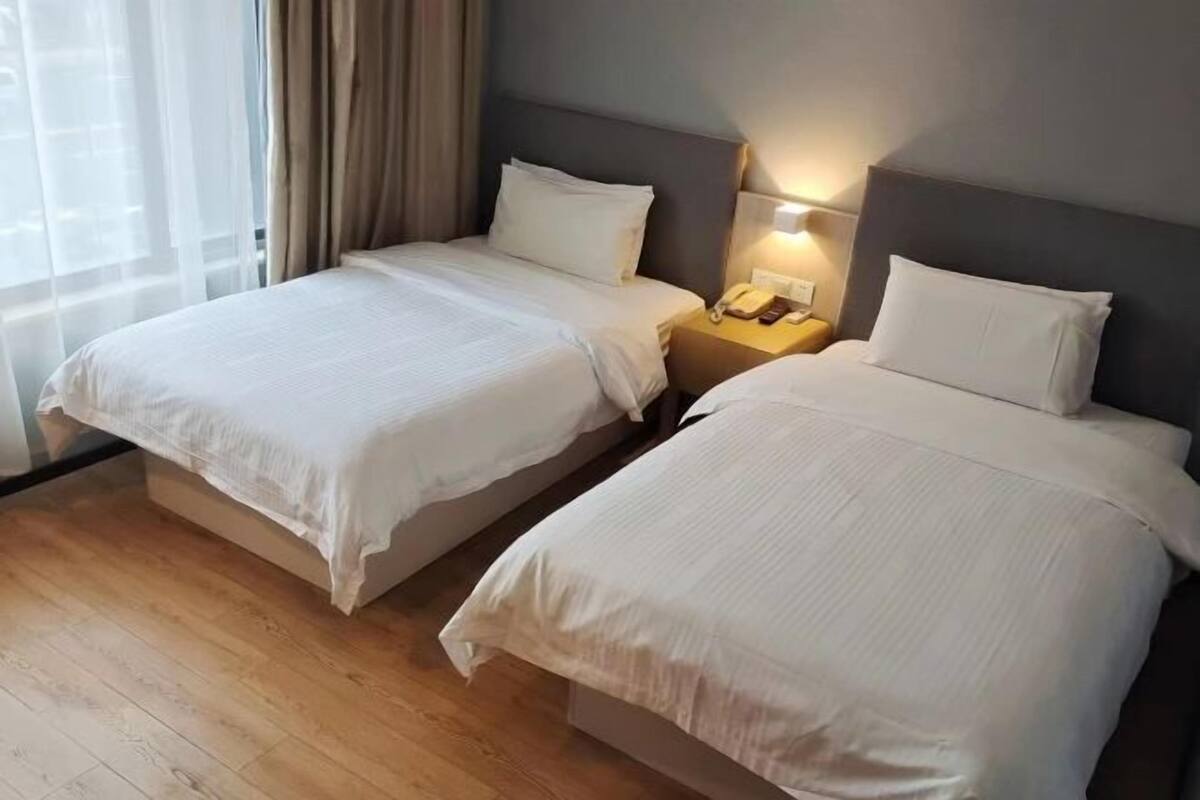 A hotel room with two beds, a bedside table with a lamp, and a window with curtains.