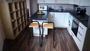 A compact kitchen with white cabinets, a microwave, a stove, and a washing machine. There are two stools and a shelf with plants. - Chelsea Orange Apartment Sleeps10-petsok-parking (London)